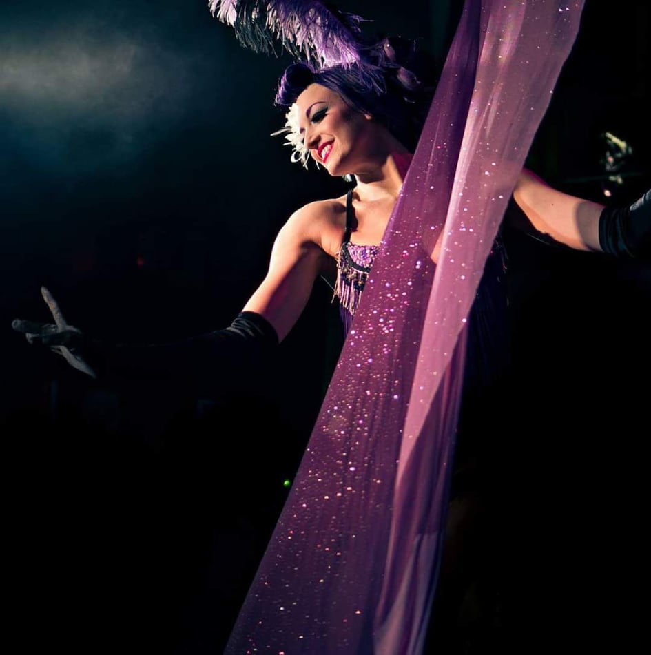 Rhapsody Blue, a white woman, onstage, sassy and bold, wears purples and black gloves, a large feather in her hair and has a large shiny fabric in front of her.