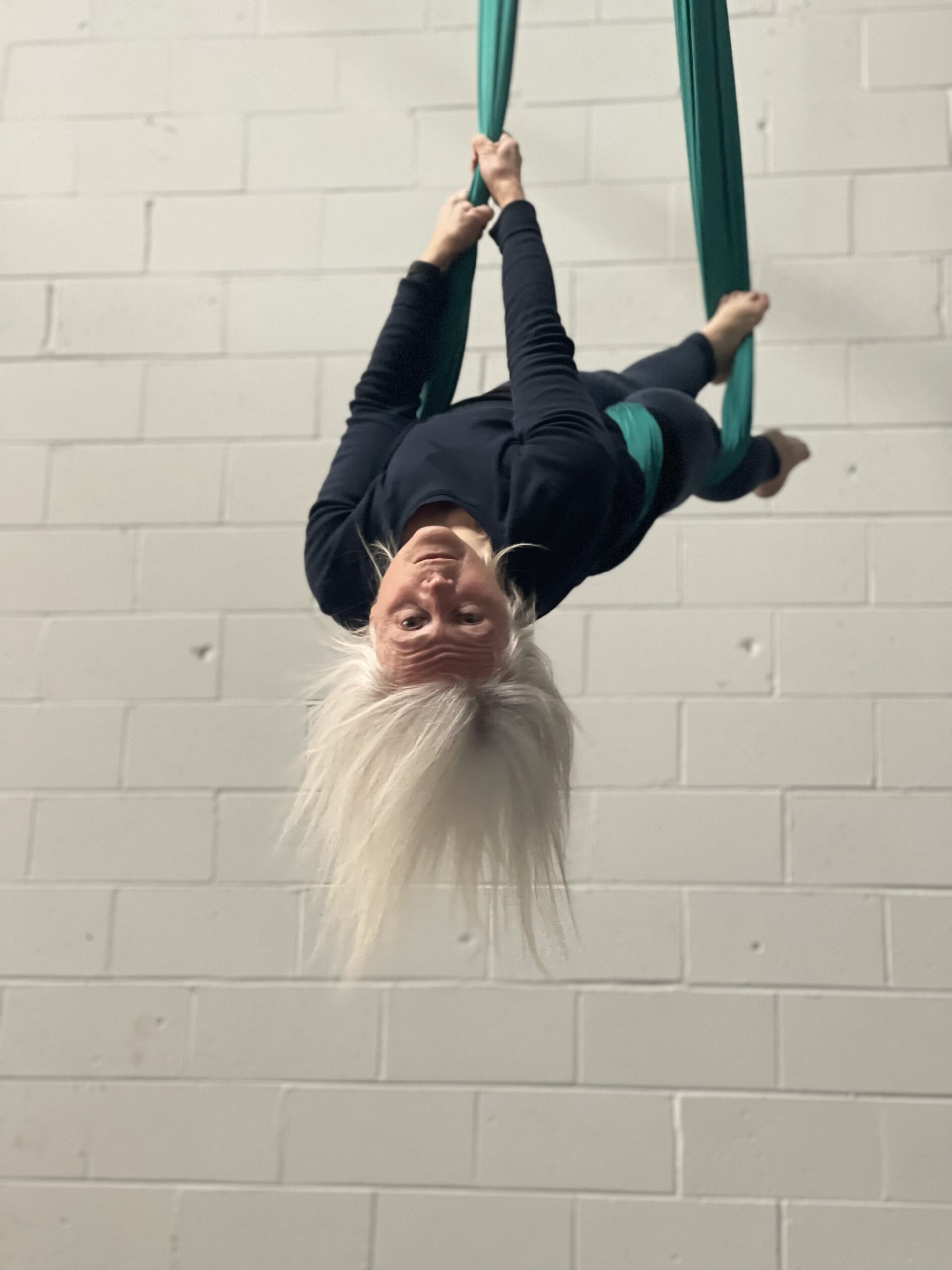 Kathleen, a white woman with silver hair, hangs upside down in the air, wrapped in aerial fabric that is suspended from the ceiling. She is strong and fierce.