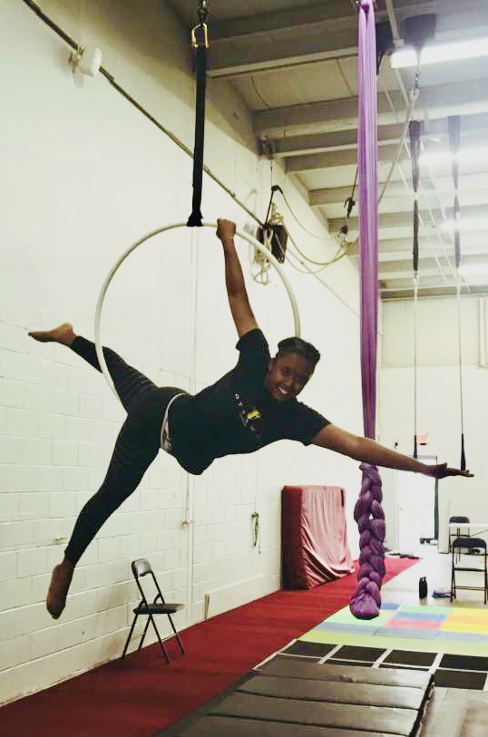 Lavie, a Black woman, hangs in the air, suspended from a large metal hoop. Her long limbs are extending and she has a huge smile that aerial arts brings to her face.